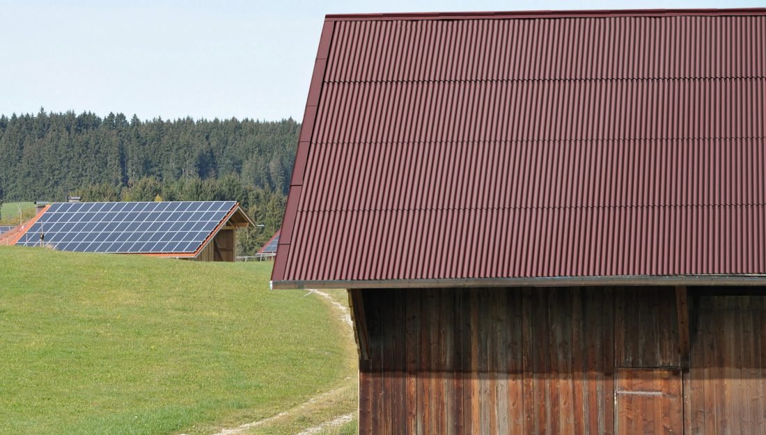 Rote Guttanit+ Bedachung auf einer Scheune vor grüner Wiese und Wald.